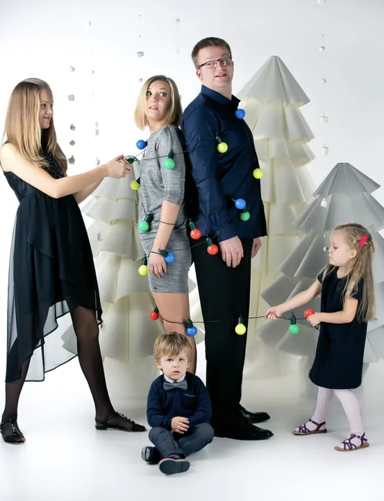 A family playfully tied together with a string of lights in front of white Christmas trees