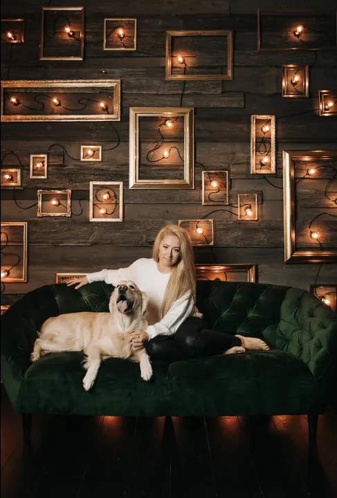 A woman and a golden retriever on a green velvet sofa with lit frames behind them